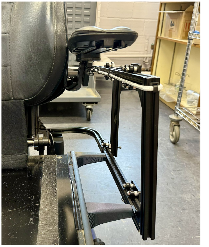 Close-up of the fabricated mechanism mounted behind the wheelchair seat, showing the extrusion rail, vertical arm, and printed supports.